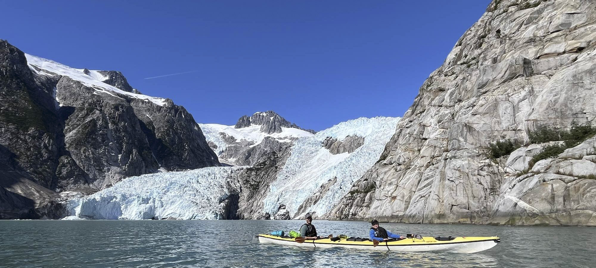 Two kayakers on turquoise water below a glacier and grey granite cliffs